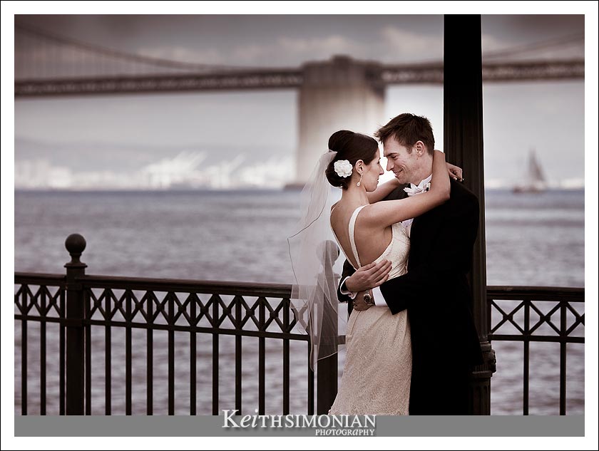 Photo showing the bride and groom on pier with Oakland - San Francisco Bay Bridge in the background