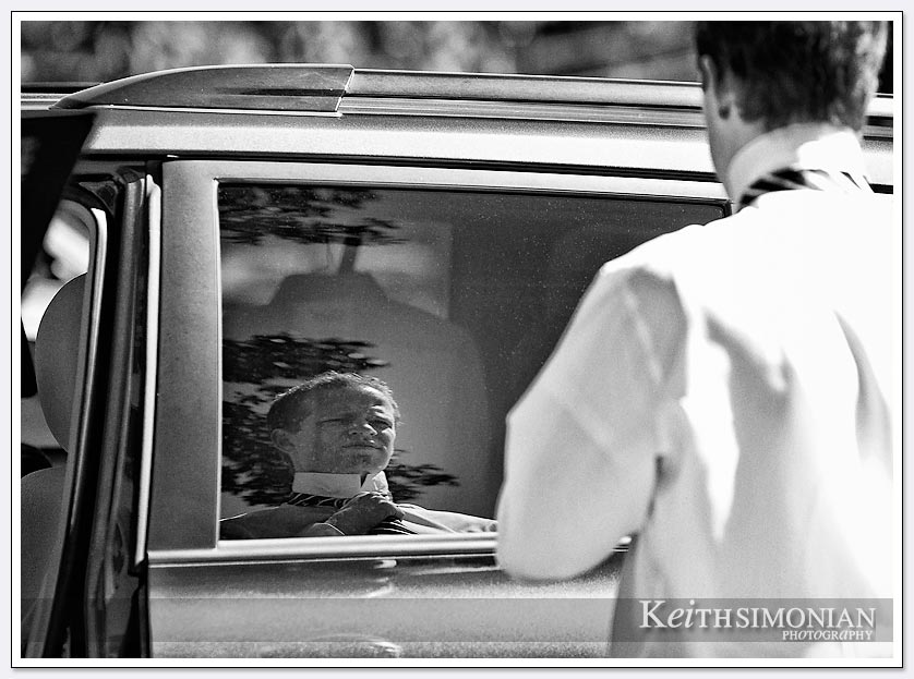 This groomsmen is using his reflection in a car window as mirror to help him tie his tie.