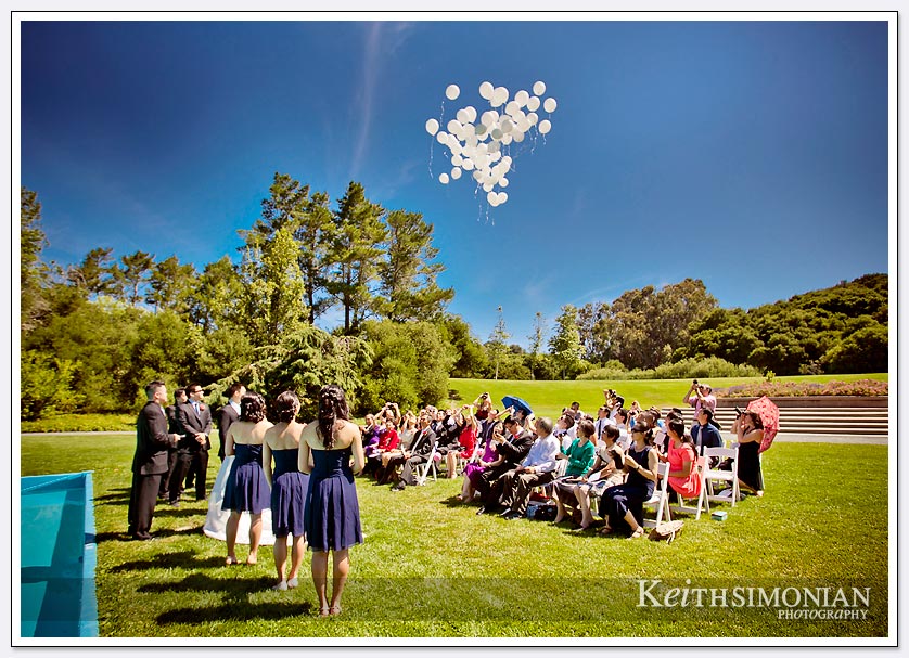 Bridal party and guests watch as white balloons are released into the blue at the end of wedding ceremony