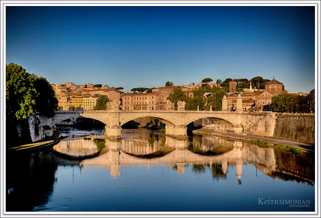The low angle of the early morning sunlight allows the reflection of the Ponte vittorio Emanuele II bridge on the blue water of the Tiber River in Rome.